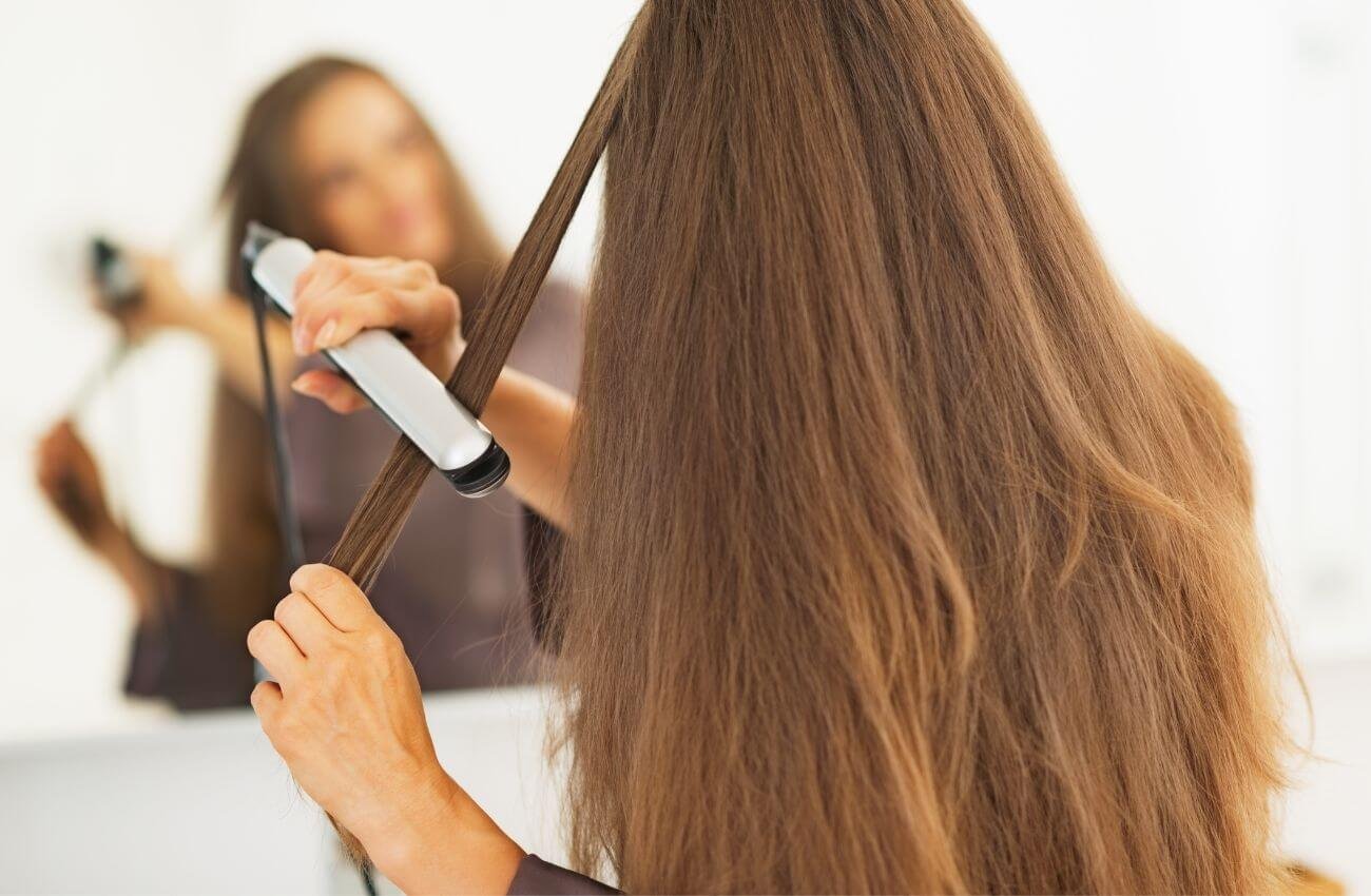 Woman Straightening Hair In Mirror - Hair Extensions Aftercare Guide